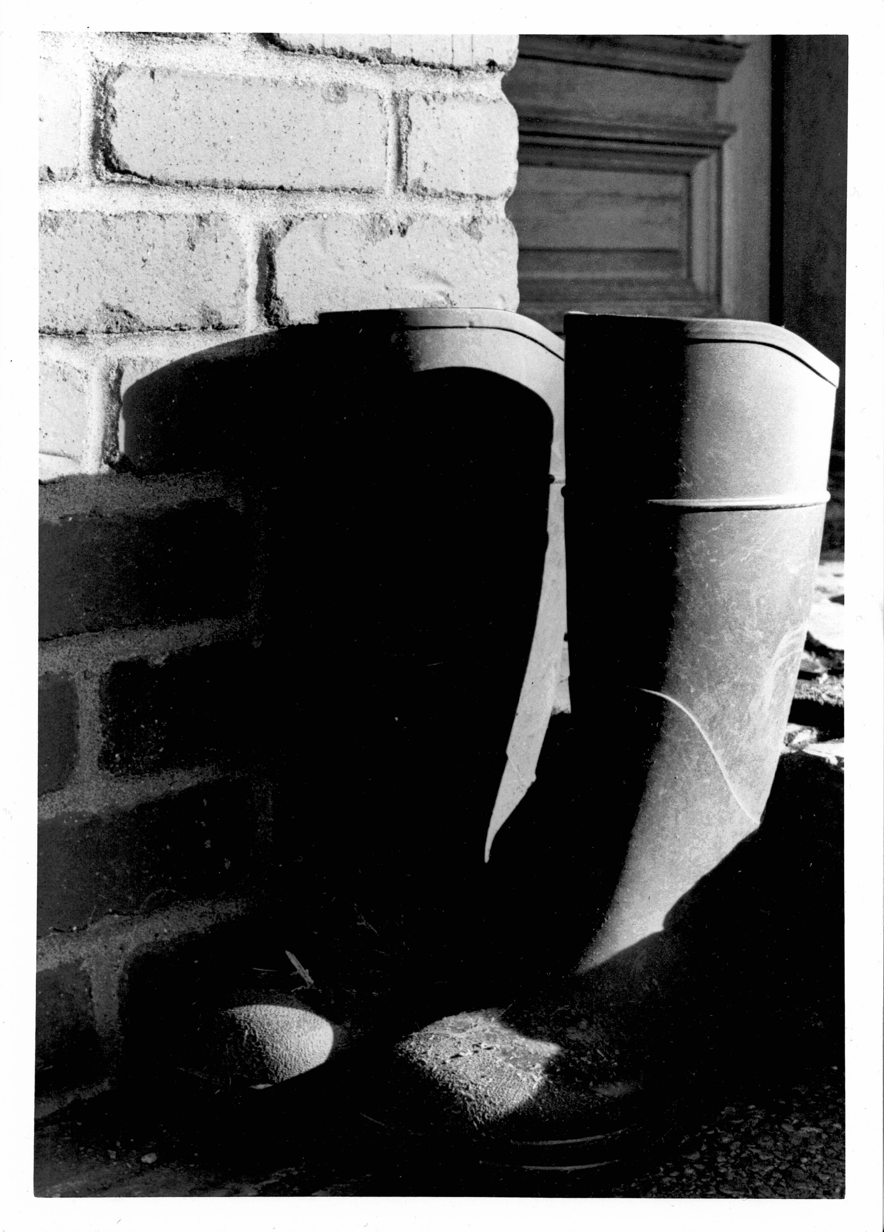 Black and white photograph. Dark shadows dance across something standing against the bricks. It's a pair of rubber work boots. Their shape is mostly obscured, but the side of one boot and the toes are brightly illuminated.