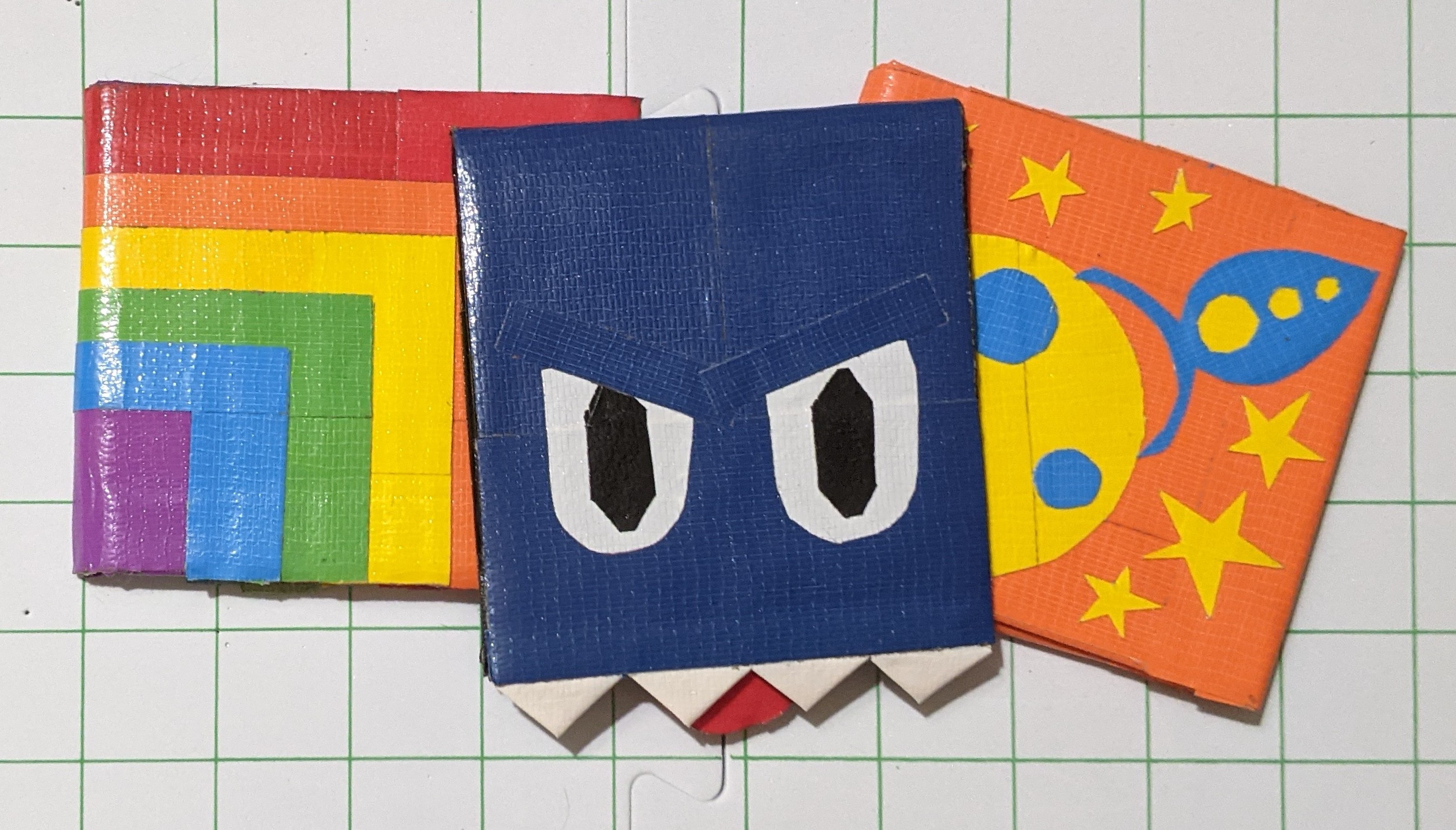 Three duct tape wallets with decorative covers: a rainbow, a rocket ship on the moon, and a grumpy monster. The opening of the wallet is its mouth.