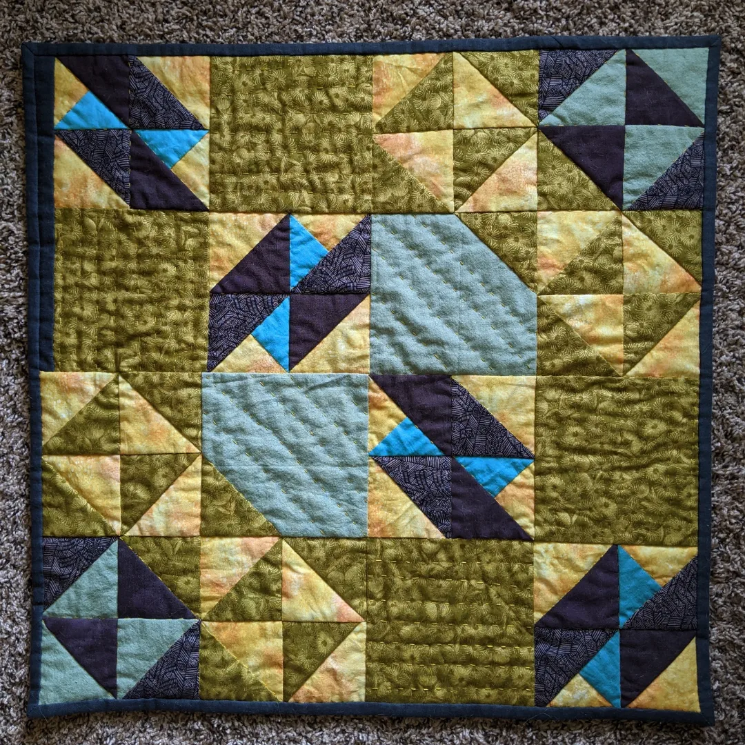 Small square quilt with muted florals. There is a line of four bright blue shapes going across the diagonal.
