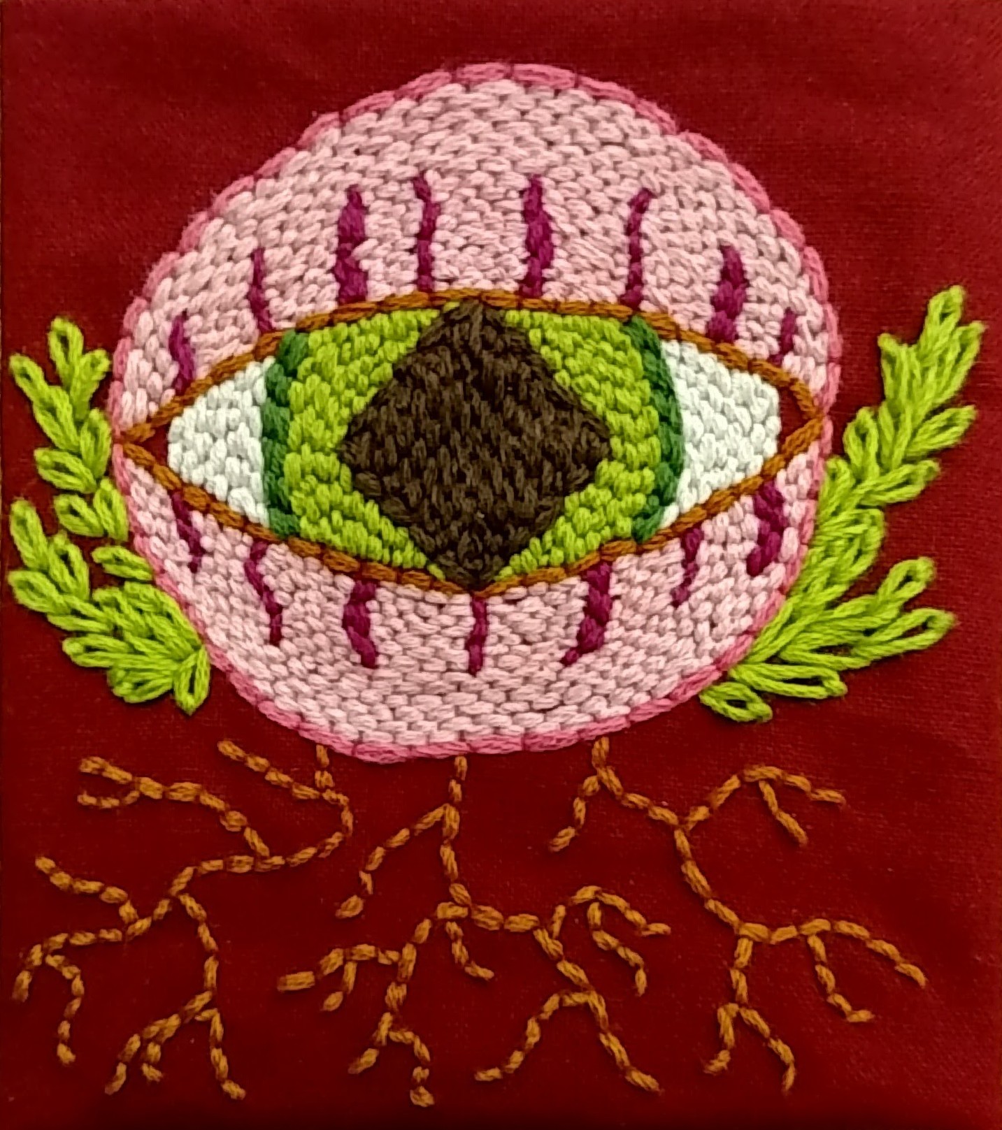 A big round eyeball with green eyes and a diamond pupil is staring straight at you, embroidered in a dense brick-like stitch pattern. Clusters of chain-stitch leaves and wandering back-stitch roots sprout from it.