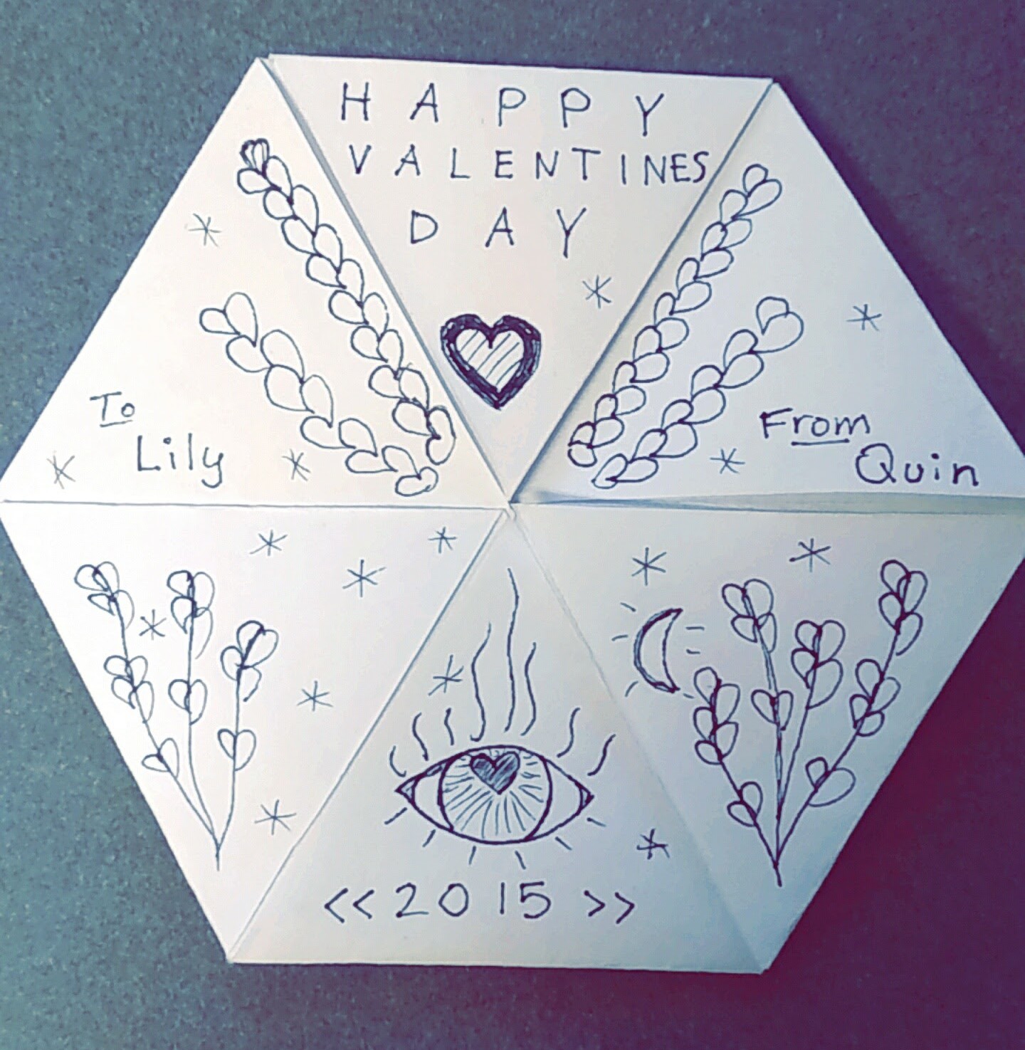 White paper folded into a hexagon, covered in pencil doodles of stars, plants, and an eye with a heart pupil. It reads: Happy valentines day. To Lily, From Quin. 2015.