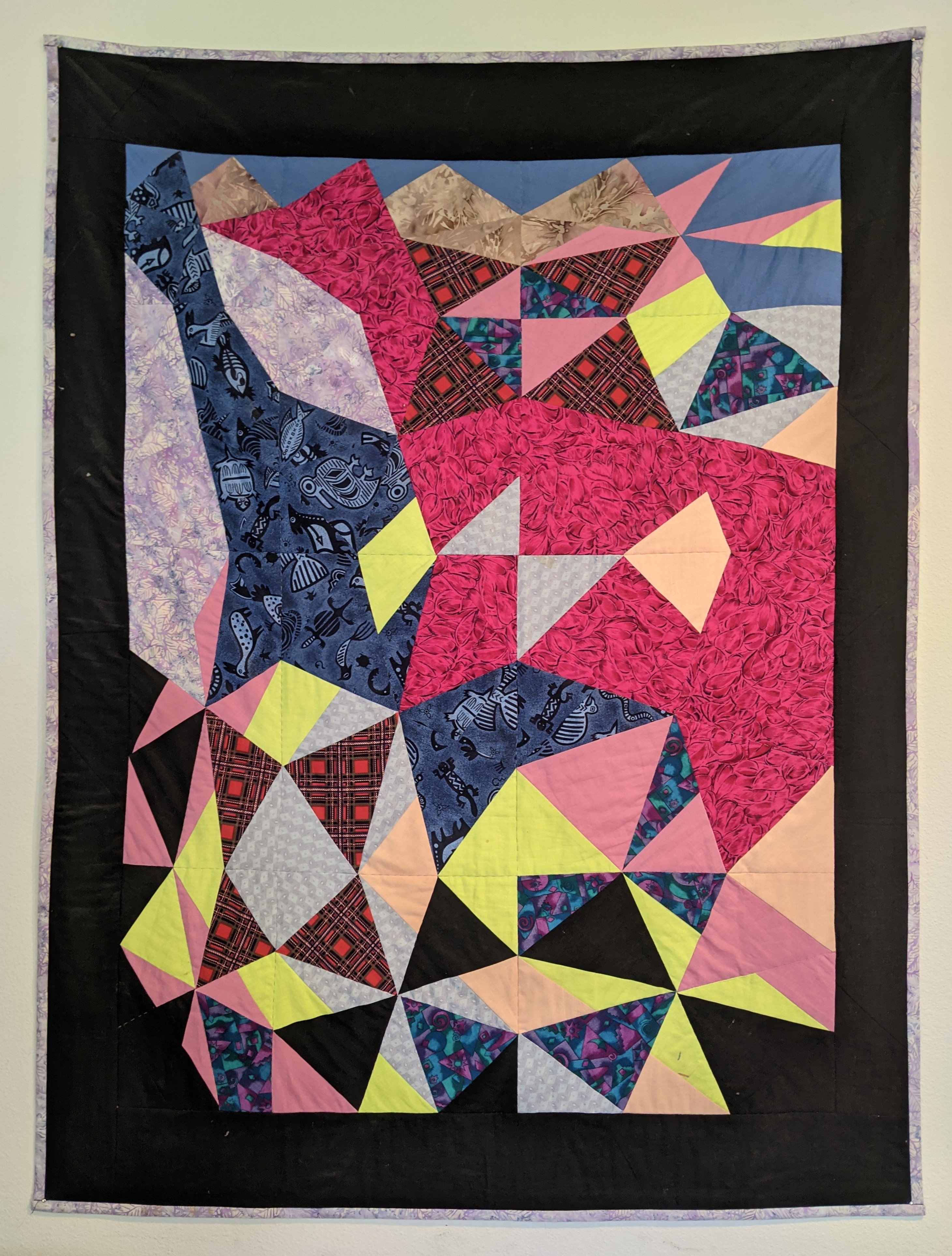 You see an abstract pink animal from above. There are brightly colored triangles flying all around. Below is the animal's shadow. It is a quilt hanging on the wall.