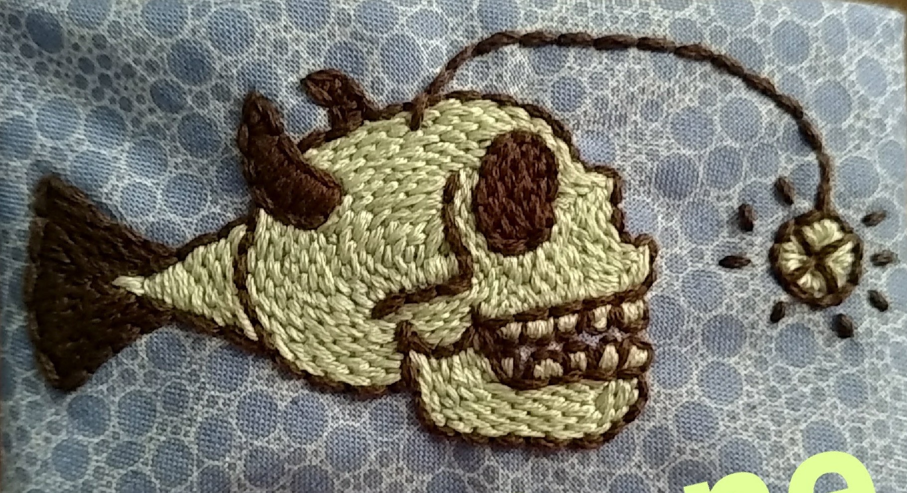 A hybrid of a human skull and an angler fish. It has horns, a fish tail, and a glowing lure that shines in front of it. It is desnsely stitched in a brick-like pattern that follows the contours of the skull, and sits against a fabric with a bubble background.
